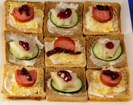A plate of open face Danish-style sandwiches, as seen from above, arranged in a checkerboard pattern. This time garnishes (lingonberry jam and sundried tomato) are visible